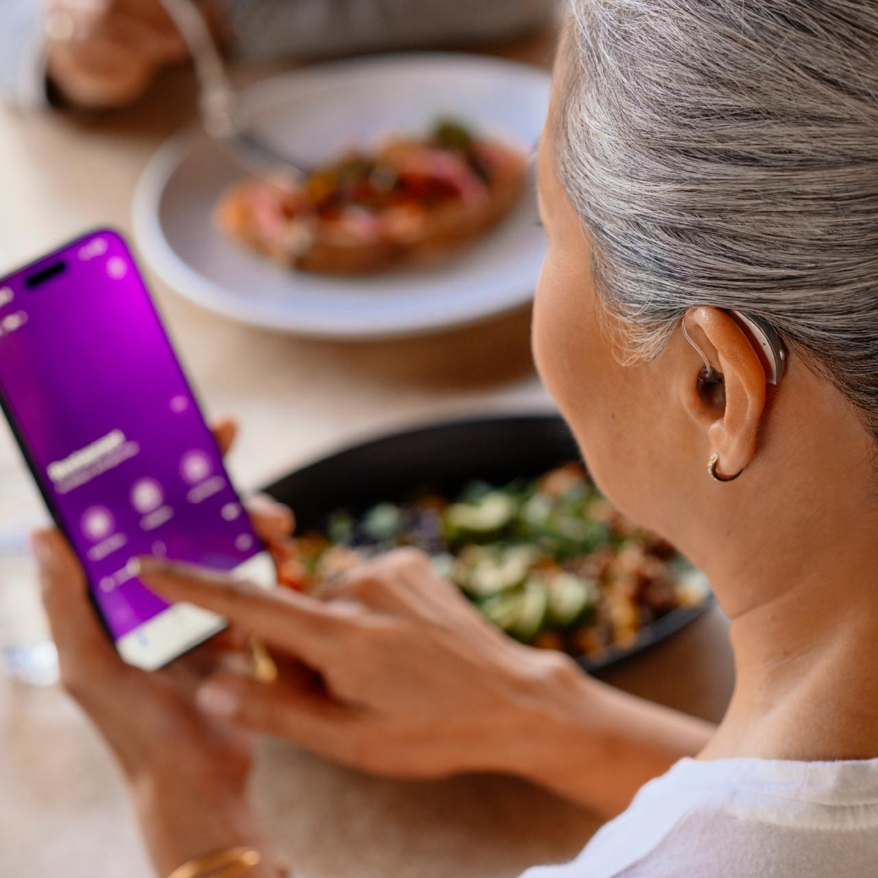 Adult woman wearing a discreet receiver-in-canal (RIC) hearing aid while controlling settings from a smartphone app at MOM's Speech & Hearing Clinic, Vijayawada